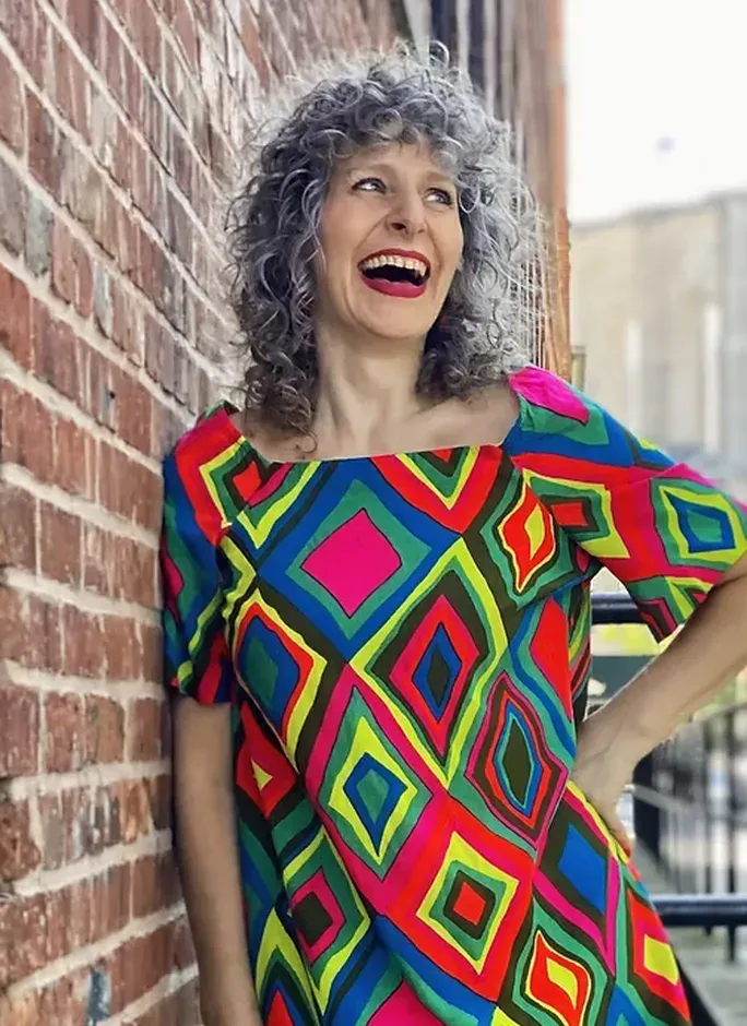 A laughing woman with curly grey hair in a brightly patterned dress, leaning against a brick wall