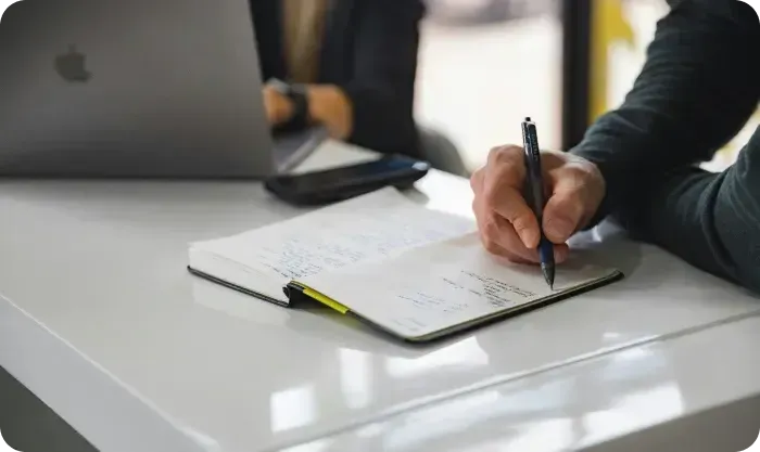Hand writing notes in a notebook on a table beside a laptop