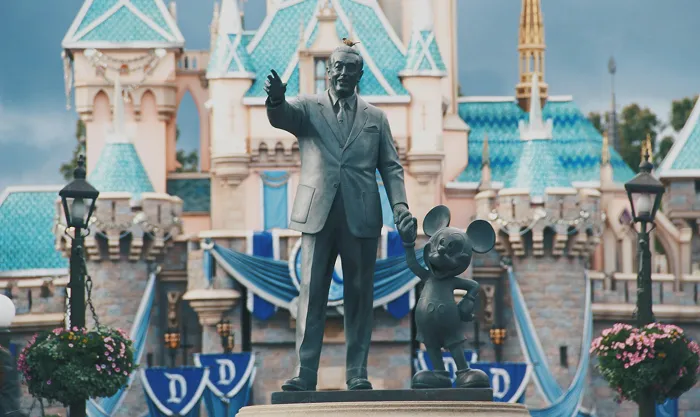 Statue of a man holding hands with a cartoon mouse in front of a fairytale castle