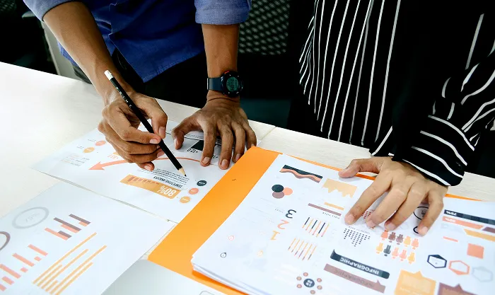 Two people pointing at printed infographics and charts on a desk