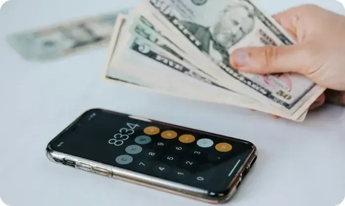 Hand holding US dollar bills above a smartphone showing a calculator
