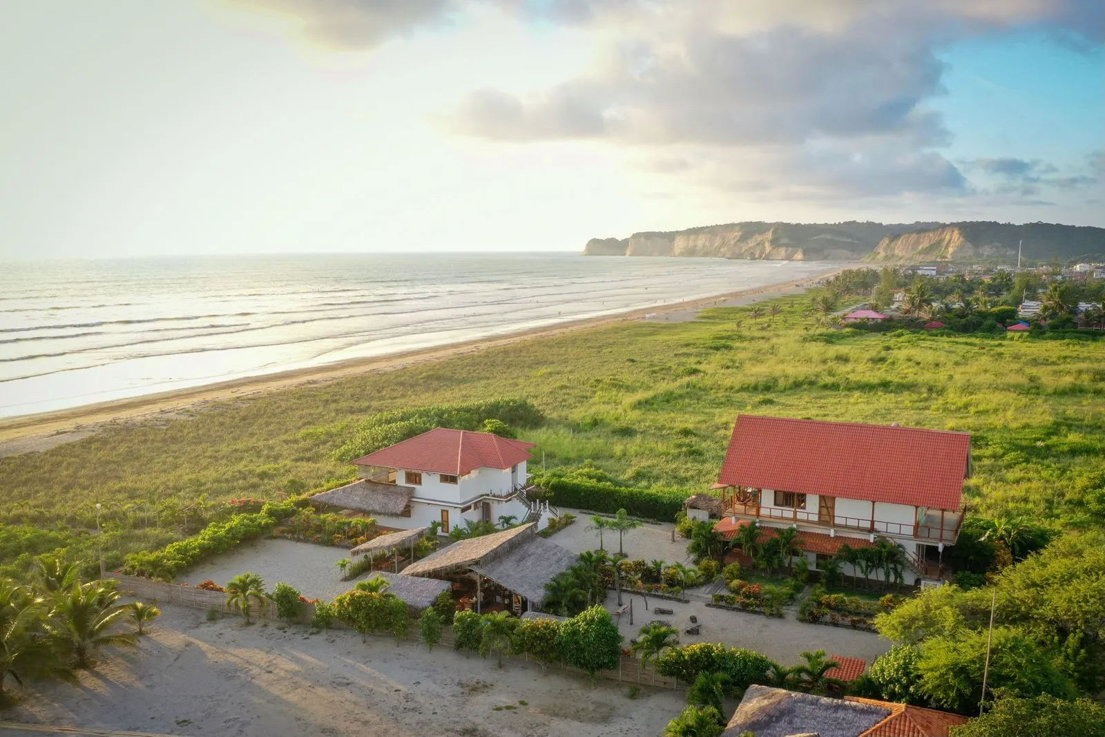 Aerial view of two red-roofed buildings among greenery beside a beach at sunset