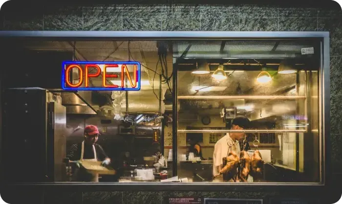Restaurant kitchen seen through a window with a neon Open sign and cooks at work