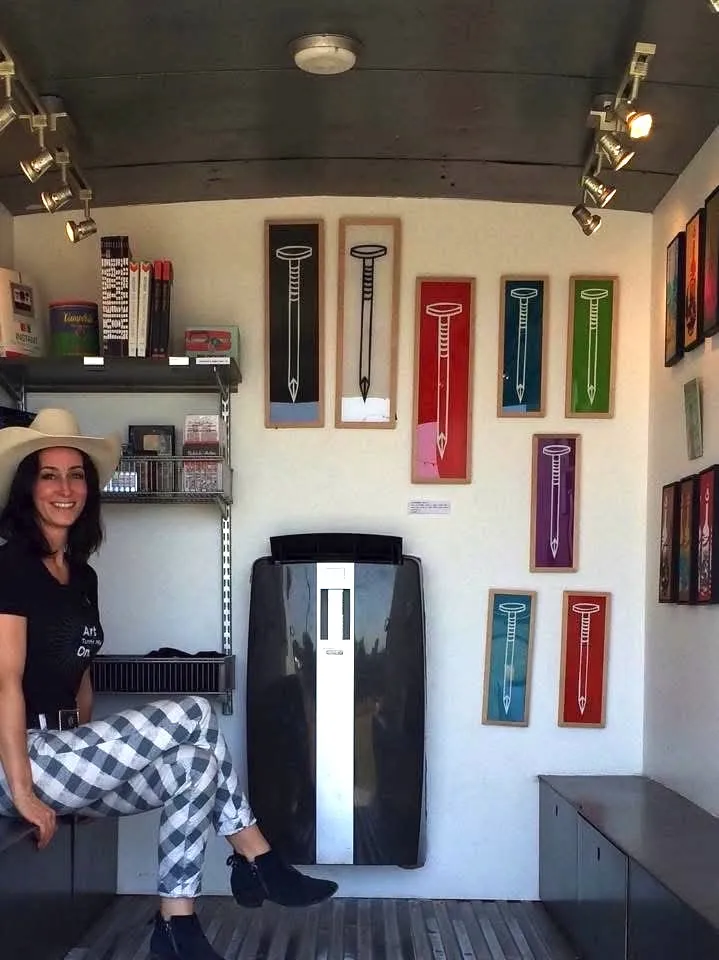 Smiling woman in a cowboy hat sitting in a small shop with framed nail illustrations on the wall