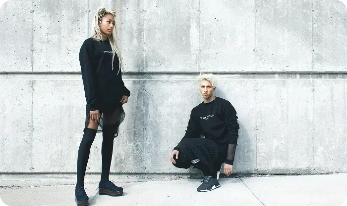 Two models in black streetwear posing against a concrete wall