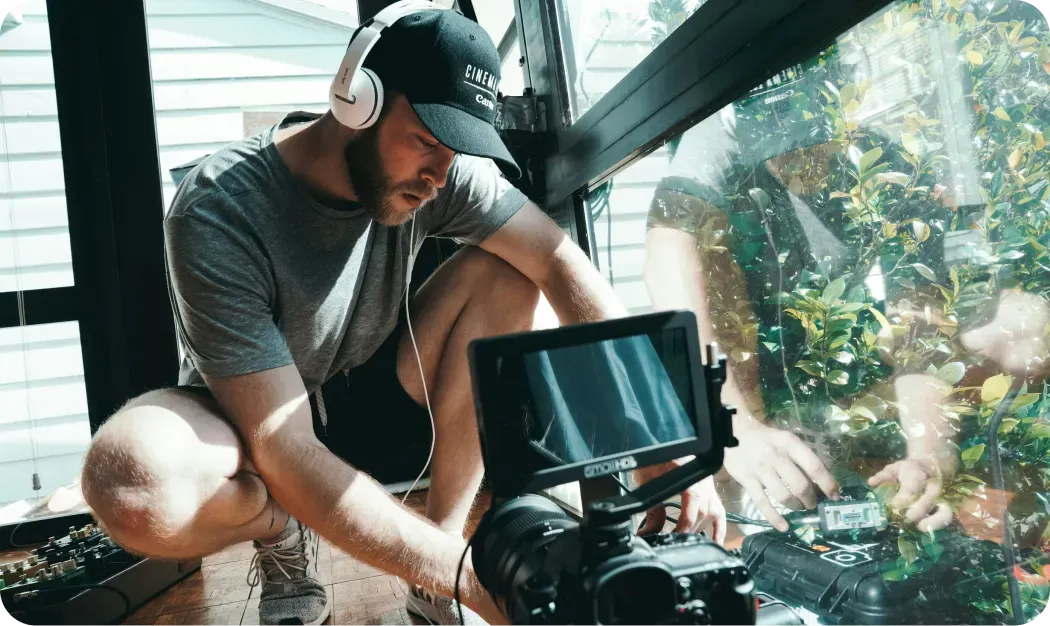 Filmmaker in headphones and a cap crouching beside a camera and audio equipment