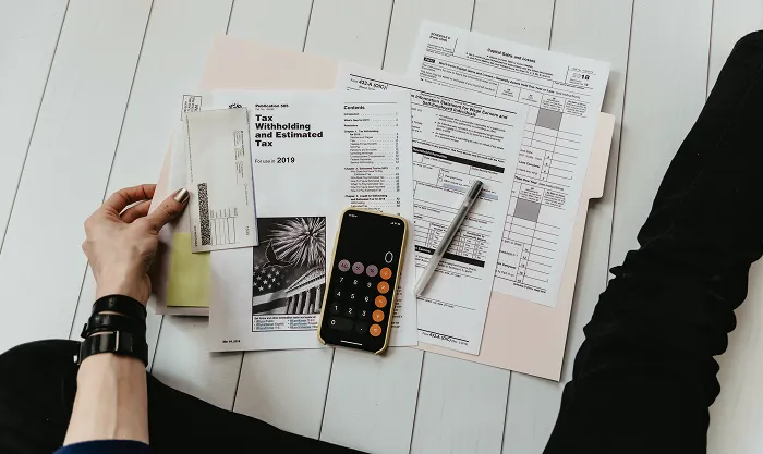 Person sitting on the floor sorting tax forms and a calculator app on a phone