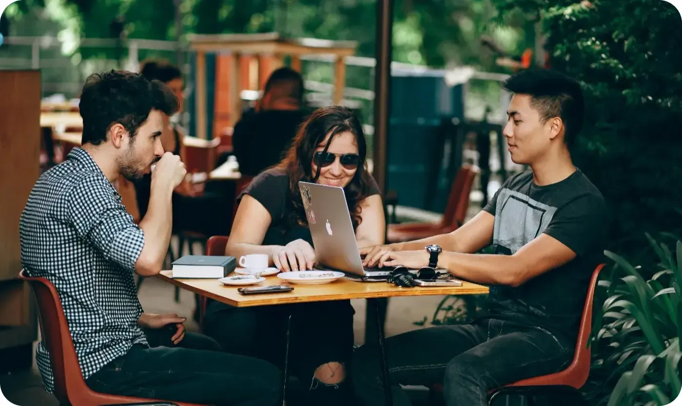 Three people sitting at an outdoor cafe table, looking at a laptop together