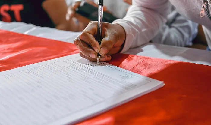 Close-up of a hand signing a printed document with a ballpoint pen on a red tablecloth