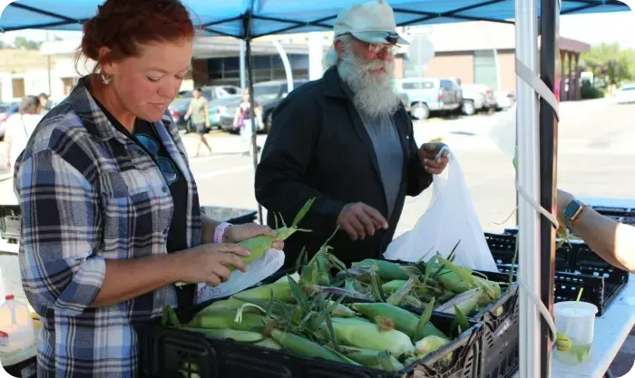 Woman at a farmers market stand bagging sweet corn for a bearded customer
