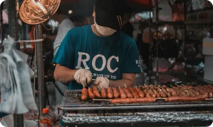 Street vendor in a mask and gloves grilling sausages on a charcoal grill