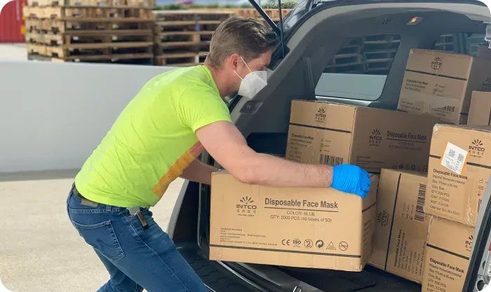 Man in a face mask and gloves loading boxes of disposable face masks into a car trunk