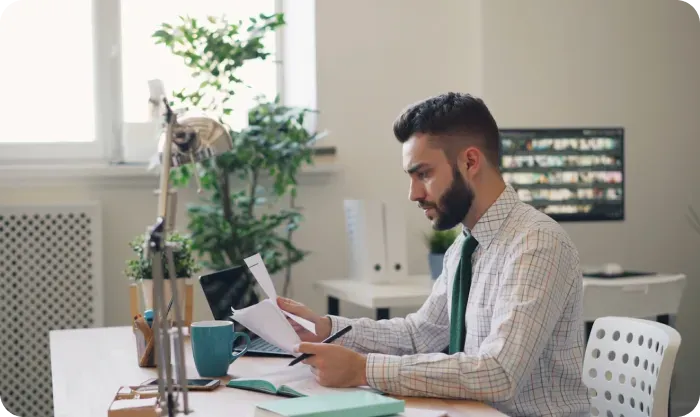 Man in a checked shirt and green tie reviewing papers at a desk with a laptop
