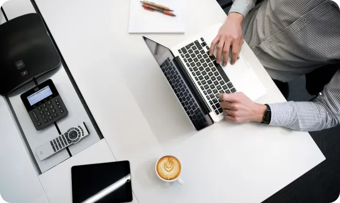 Overhead view of a man typing on a laptop beside a latte, tablet and desk phone