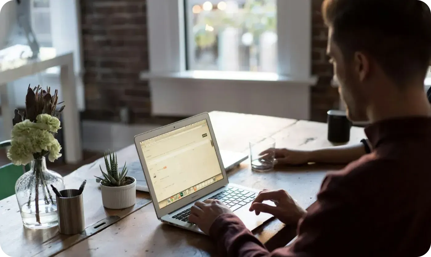 Man typing on a laptop at a wooden table with a succulent and flowers