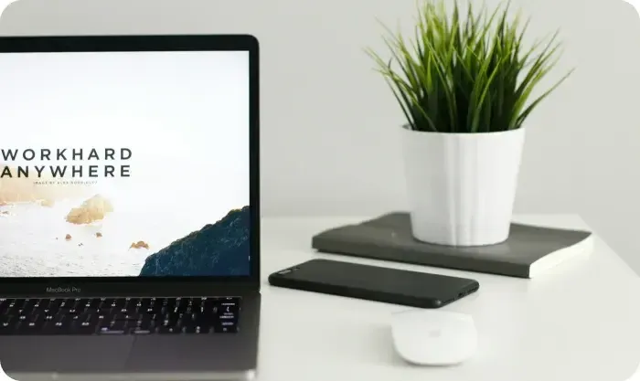 Laptop showing the words Work Hard Anywhere beside a potted plant, phone and mouse on a white desk
