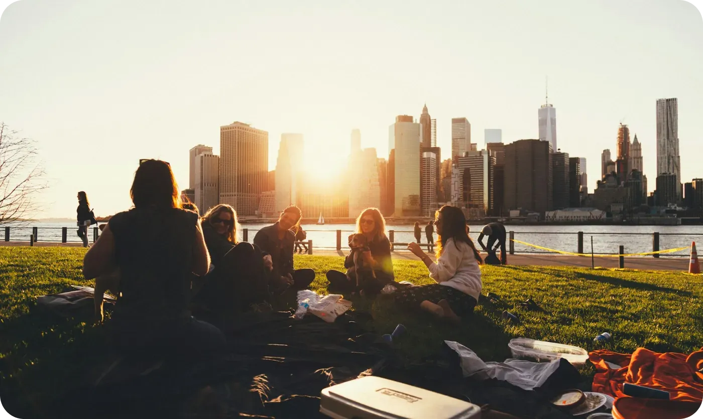 Friends sitting on a grassy waterfront lawn at sunset with a city skyline behind them