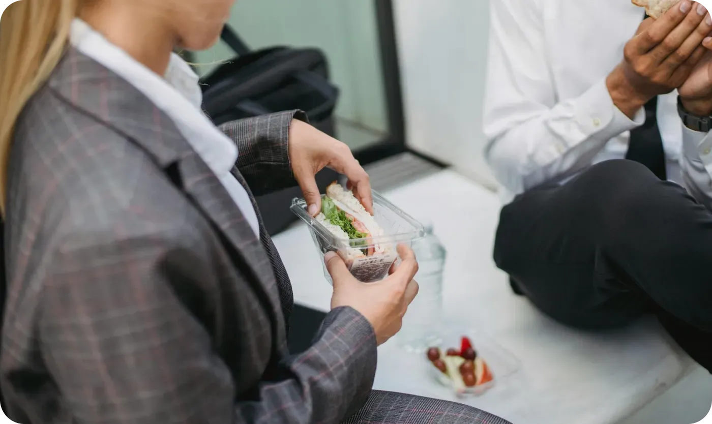 Two colleagues in business attire eating sandwiches and fruit during a lunch break