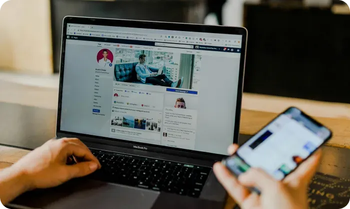 Person using a laptop showing a social media profile page while holding a smartphone