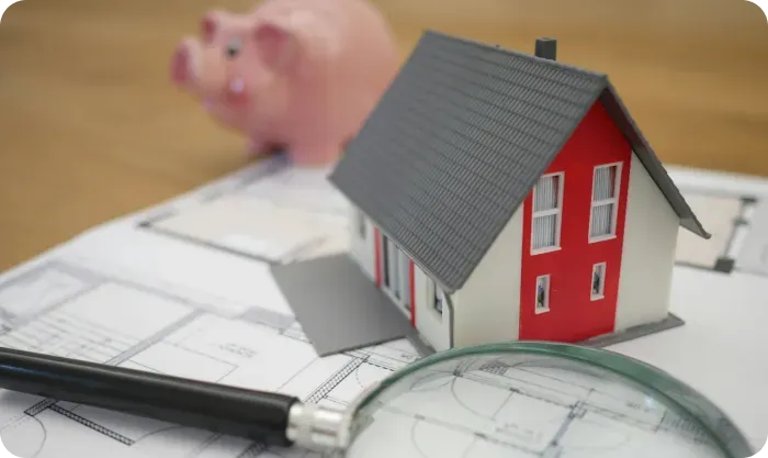 Small model house on blueprints with a magnifying glass and a piggy bank behind it
