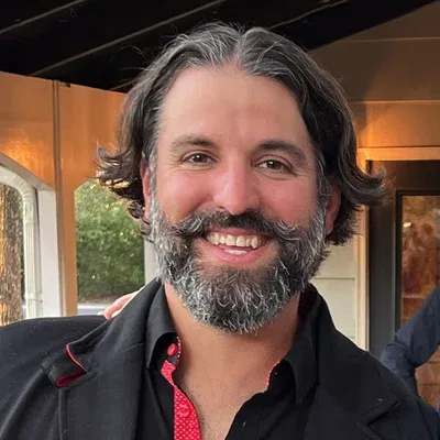 Smiling bearded man in a dark jacket and red-patterned shirt