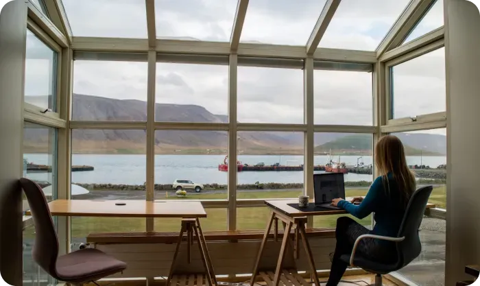 Woman working on a laptop at a wooden desk in a glass room overlooking a harbor and mountains