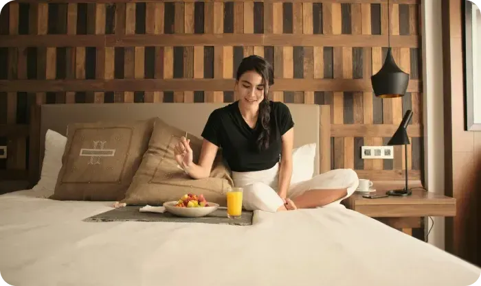 Woman eating fruit and orange juice from a breakfast tray while sitting on a hotel bed