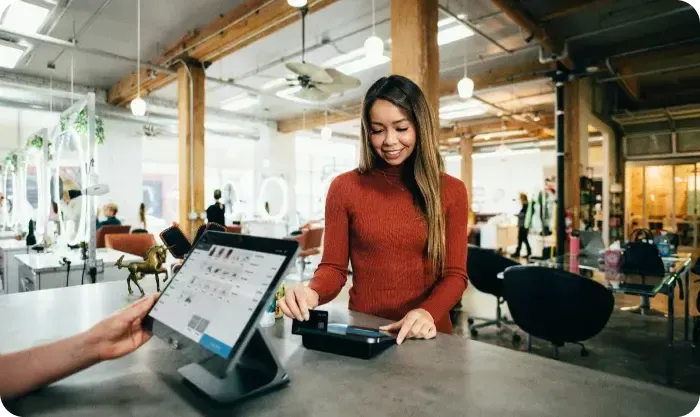 Smiling woman tapping a card on a payment reader at a counter beside a tablet register