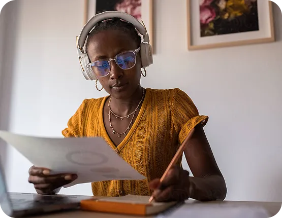 Woman wearing headphones and glasses reviewing a printed document while taking notes