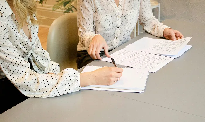Two women at a table going over a printed document, one taking notes while the other points at the text