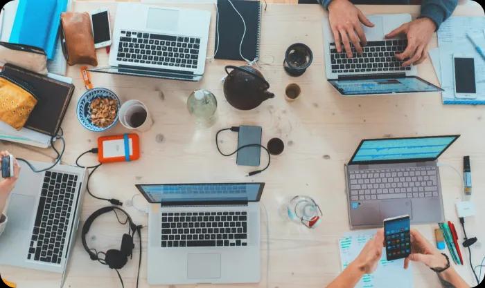 A desk with multiple laptops and accessories and four people working.