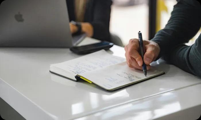 A man writing notes in a notebook and woman working on PC 