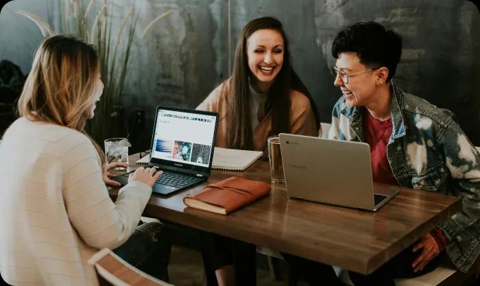 Three women engaged in discussion while working on laptops at a table, showcasing collaboration and productivity.