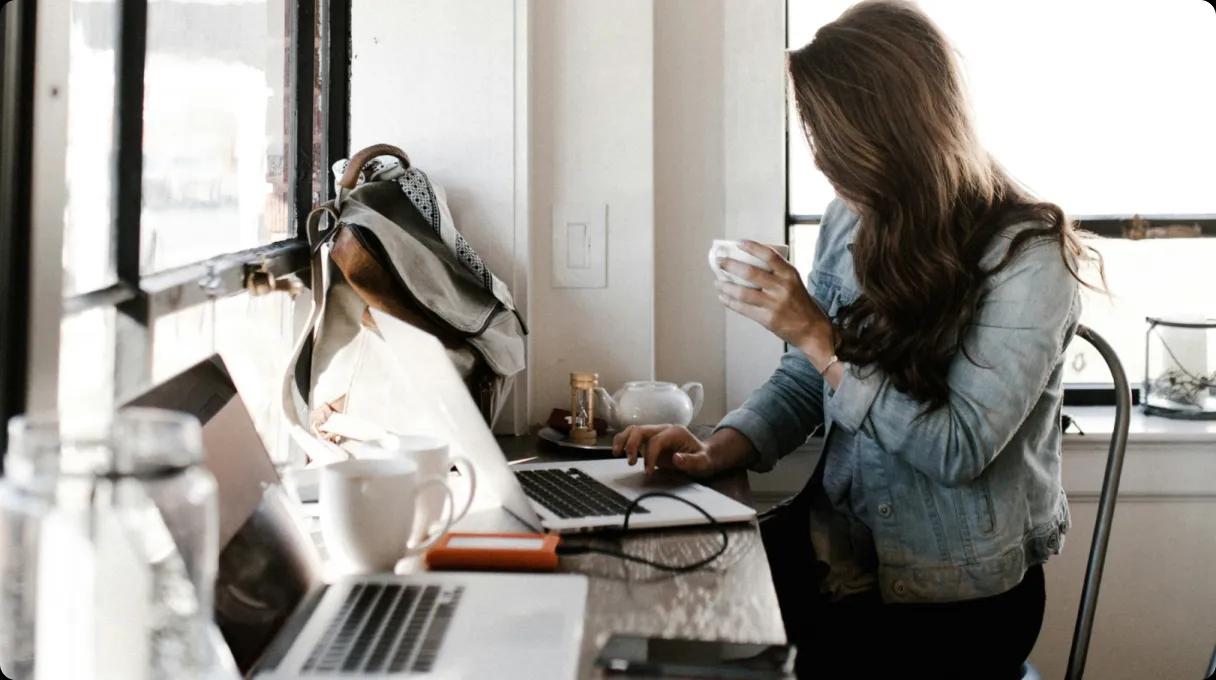 A woman holding a cup sitting in front of a desk with laptops on it, she appears to be browsing or reading.