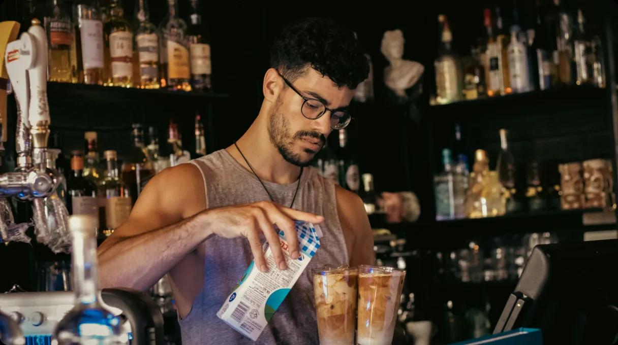 A bartender in glasses is seen pouring milk into a coffee or drink