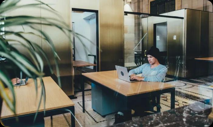 A man seated at a desk, focused on his laptop, surrounded by a tidy workspace and natural light.