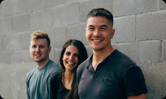  Three individuals smiling joyfully in front of a textured brick wall, showcasing a moment of happiness and camaraderie.