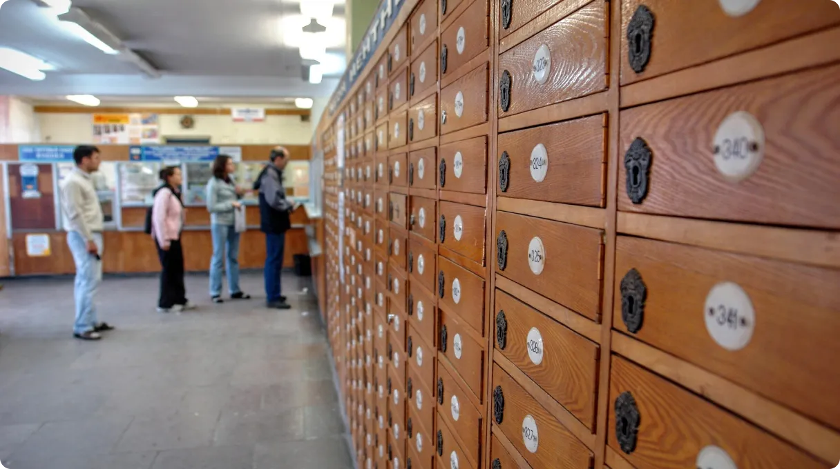 A row of wooden post office PO boxes with numbered locks, extending into the distance. In the background, a group of people stand in line at the counter inside a postal service office.