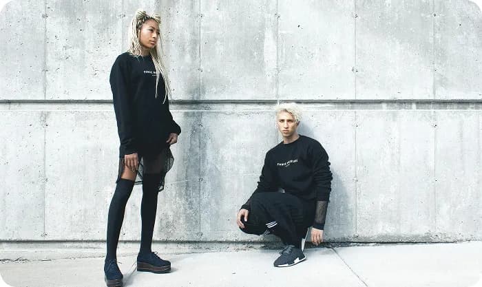 Two fashion models in black streetwear posing against a minimalist concrete wall, showcasing edgy urban style with bold aesthetics.
