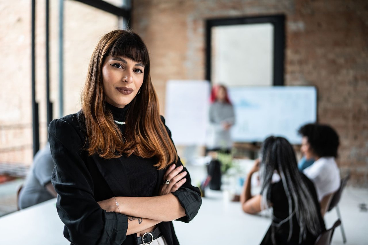 Confident female business leader standing with arms crossed in modern meeting room with team in the background.