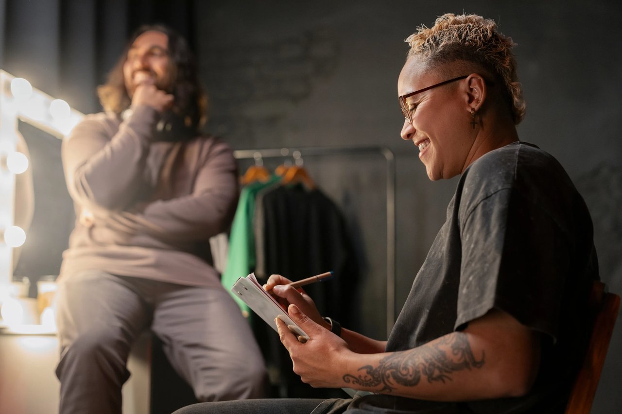 Two entrepreneurs laughing together in a creative studio, one reviewing notes on a tablet while the other leans back smiling, with a clothing rack and studio lighting in the background.