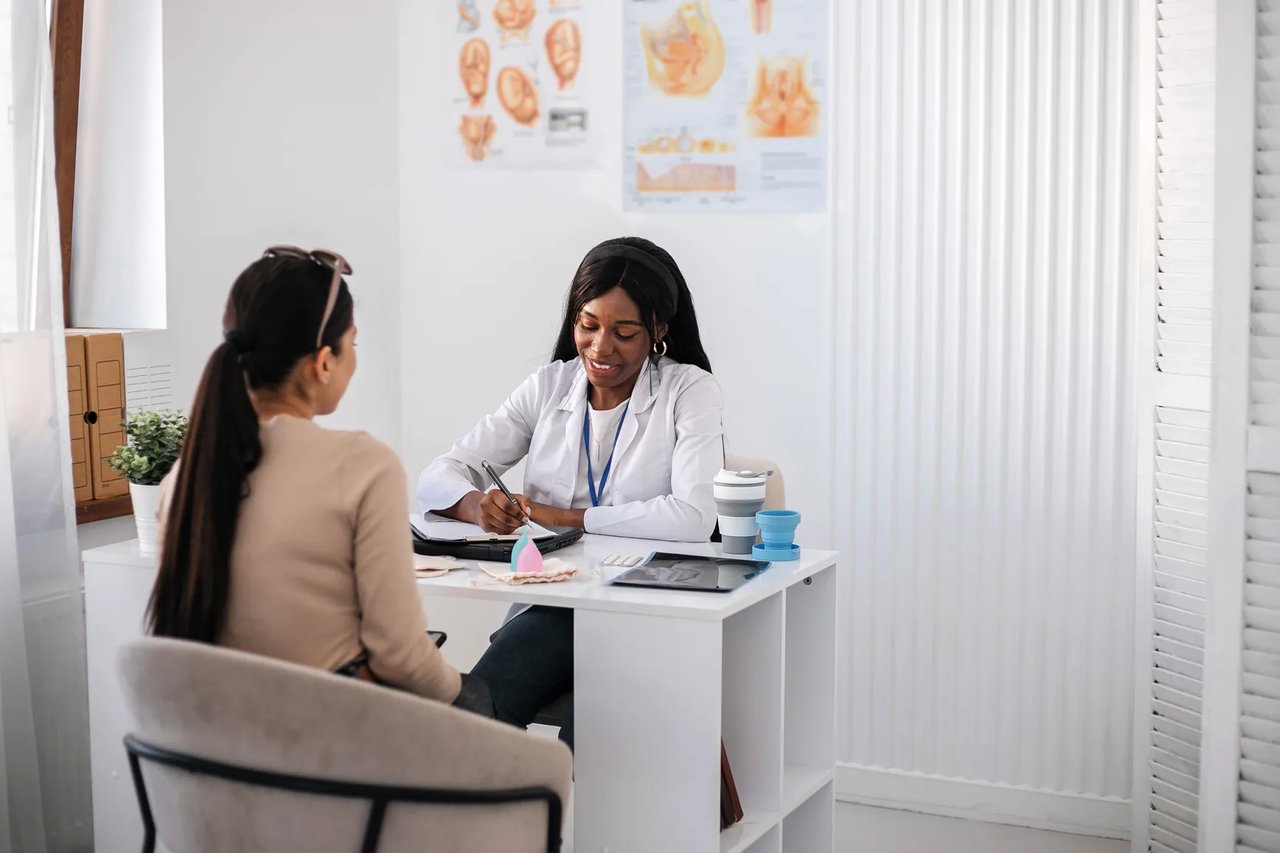 A dentist in scrubs standing at the reception desk of a small practice, reviewing paperwork, with treatment rooms visible through an open doorway behind her.