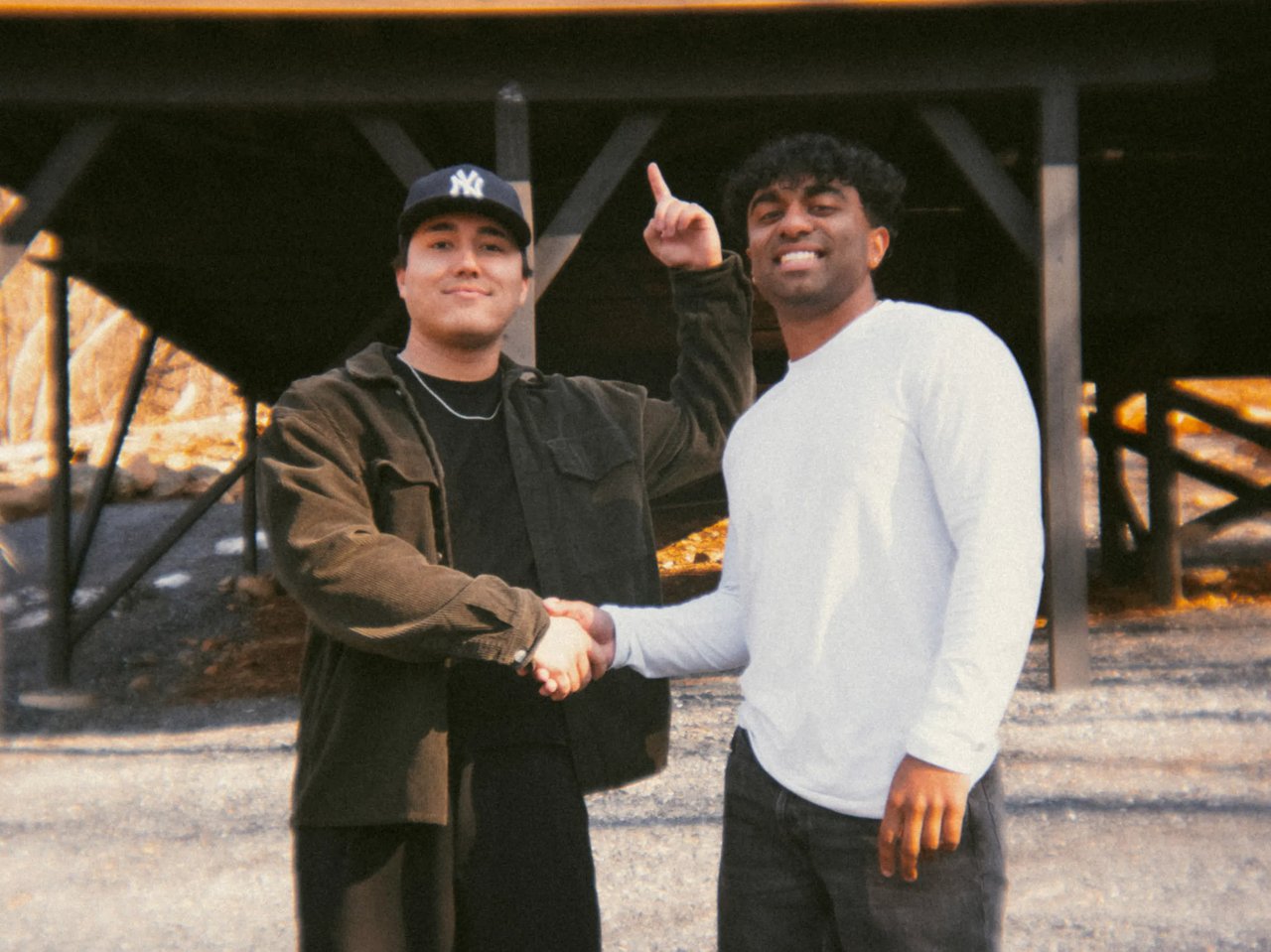 Two young men shaking hands and smiling outdoors, one in a dark jacket and NY baseball cap, the other in a white long-sleeve shirt pointing upward, standing on gravel in front of a wooden structure.