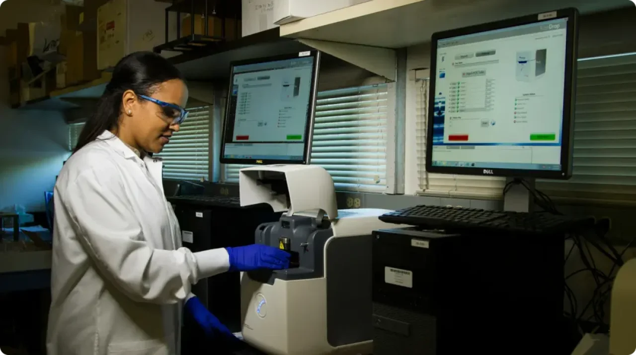 female-scientist-in-a-lab-coat-and-safety-glasses-using-a-laboratory-machine-with-computer-screens-displaying-experiment-data