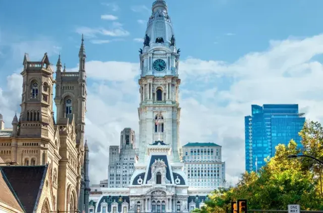 Philadelphia City Hall clock tower with surrounding historic and modern buildings under a partly cloudy sky.