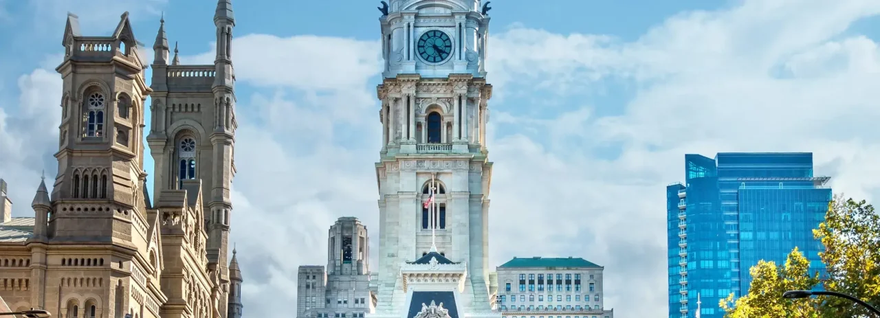 Philadelphia City Hall clock tower with surrounding historic and modern buildings under a partly cloudy sky.